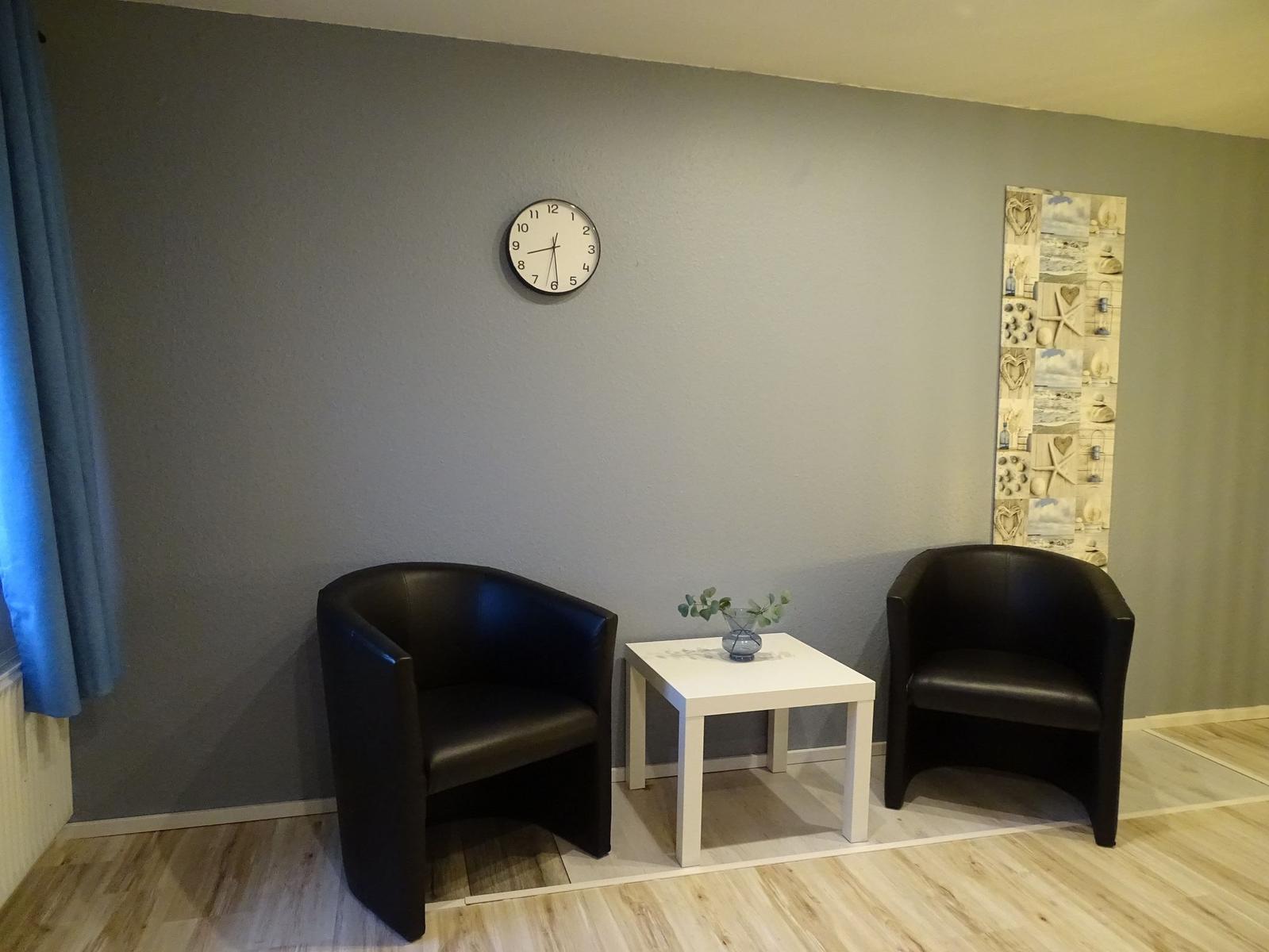 Zwei schwarze Sessel und ein weißer Tisch mit Blumen vor einer Wand mit Uhr und Wandbild.