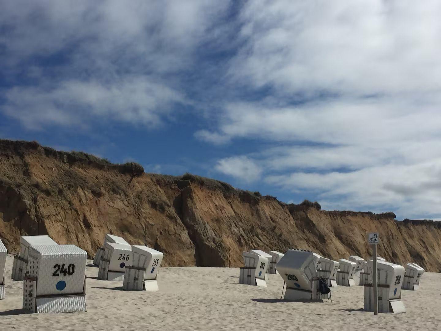 Strand mit weißen Strandkörben und sandigem Ufer vor steilen Klippen.