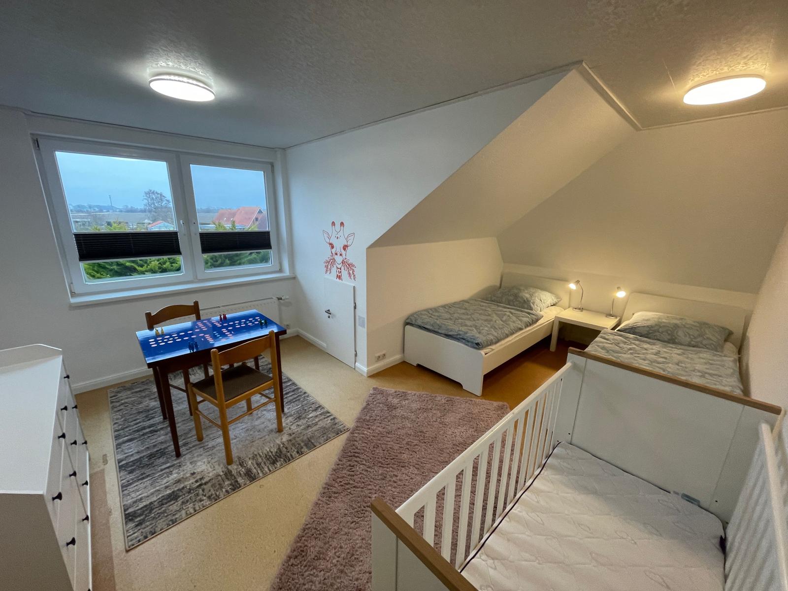 Zimmer mit zwei Betten, Kinderbett und Spielplatz mit Tisch.