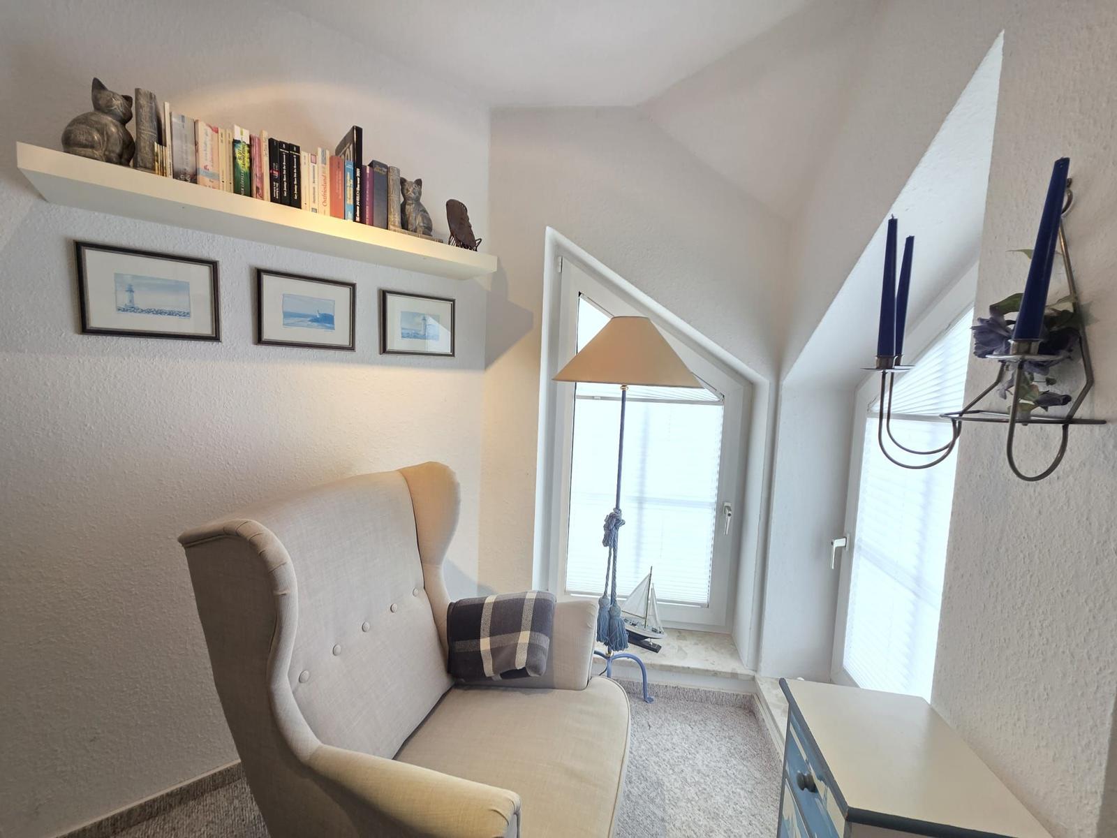 A room with a chair, windows, and a wall shelf with books and decor.