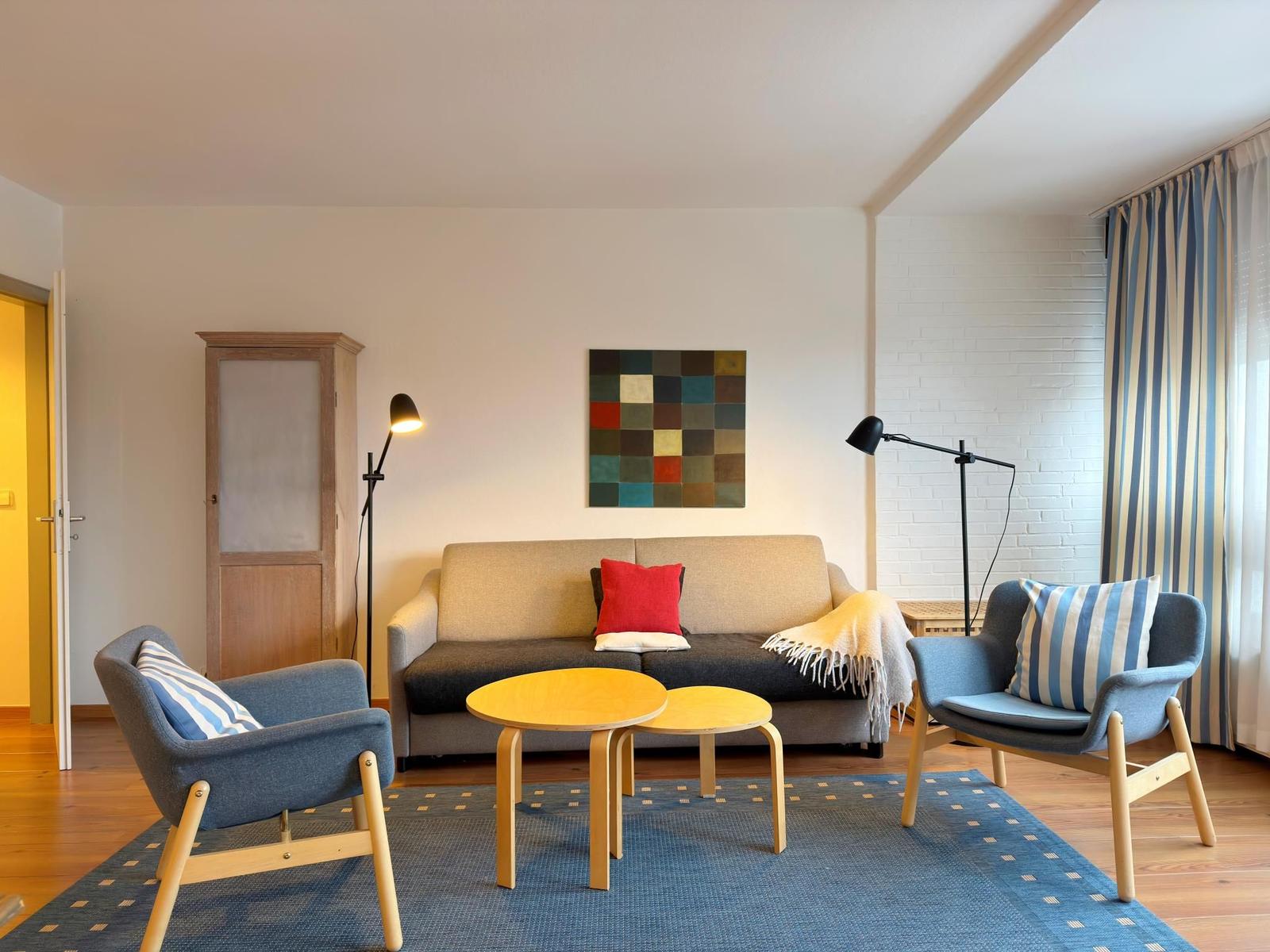 Wohnzimmer mit Sofa, zwei Sesseln und Tisch. Wandbild und Streifenstore.