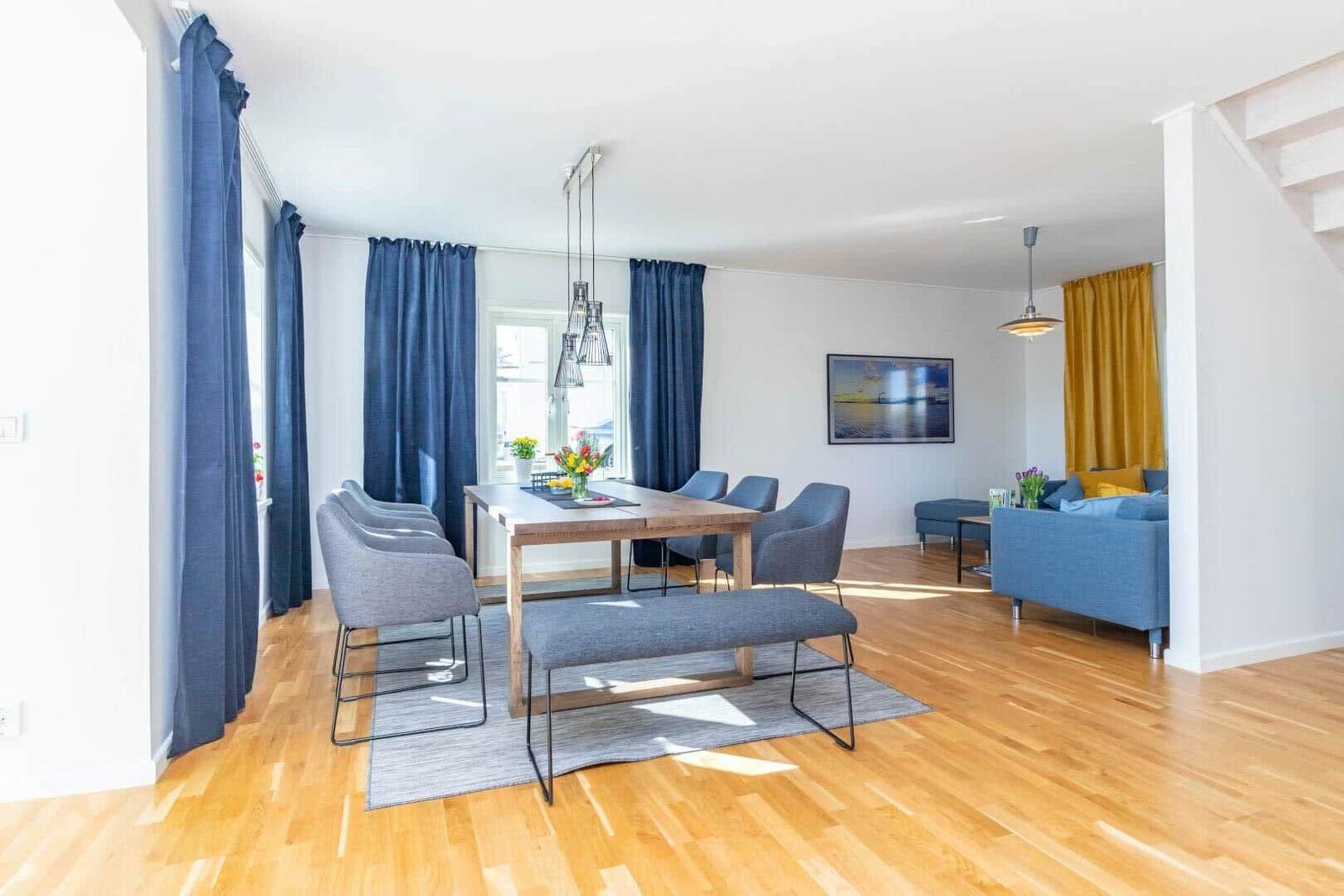 Esszimmer mit Holztisch, grauen Stühlen und blauen Vorhängen. Wohnbereich mit blauem Sofa und gelben Vorhängen.