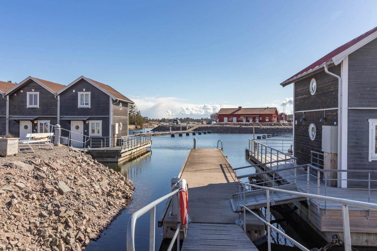 Holzhäuser am Wasser mit Steg und rotem Bootshaus im Hintergrund.