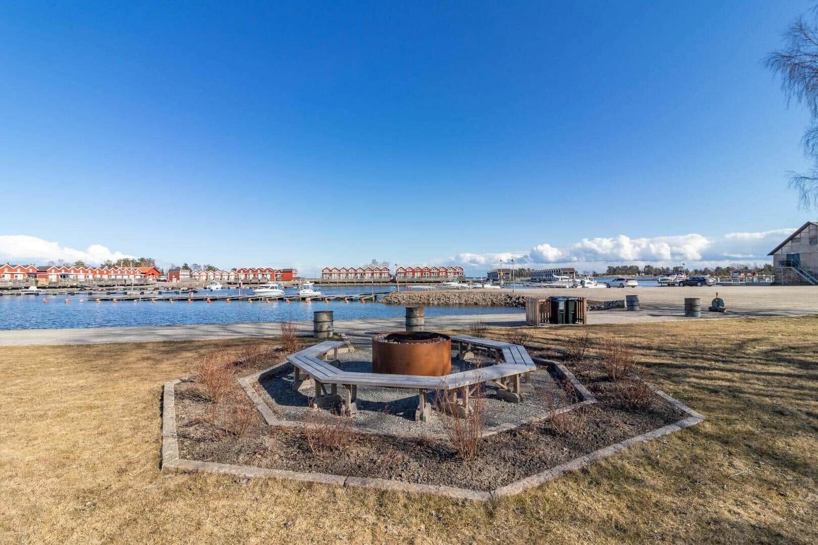 Feuerstelle mit Sitzbank am Wasser, Hintergrund mit Booten und roten Häusern.
