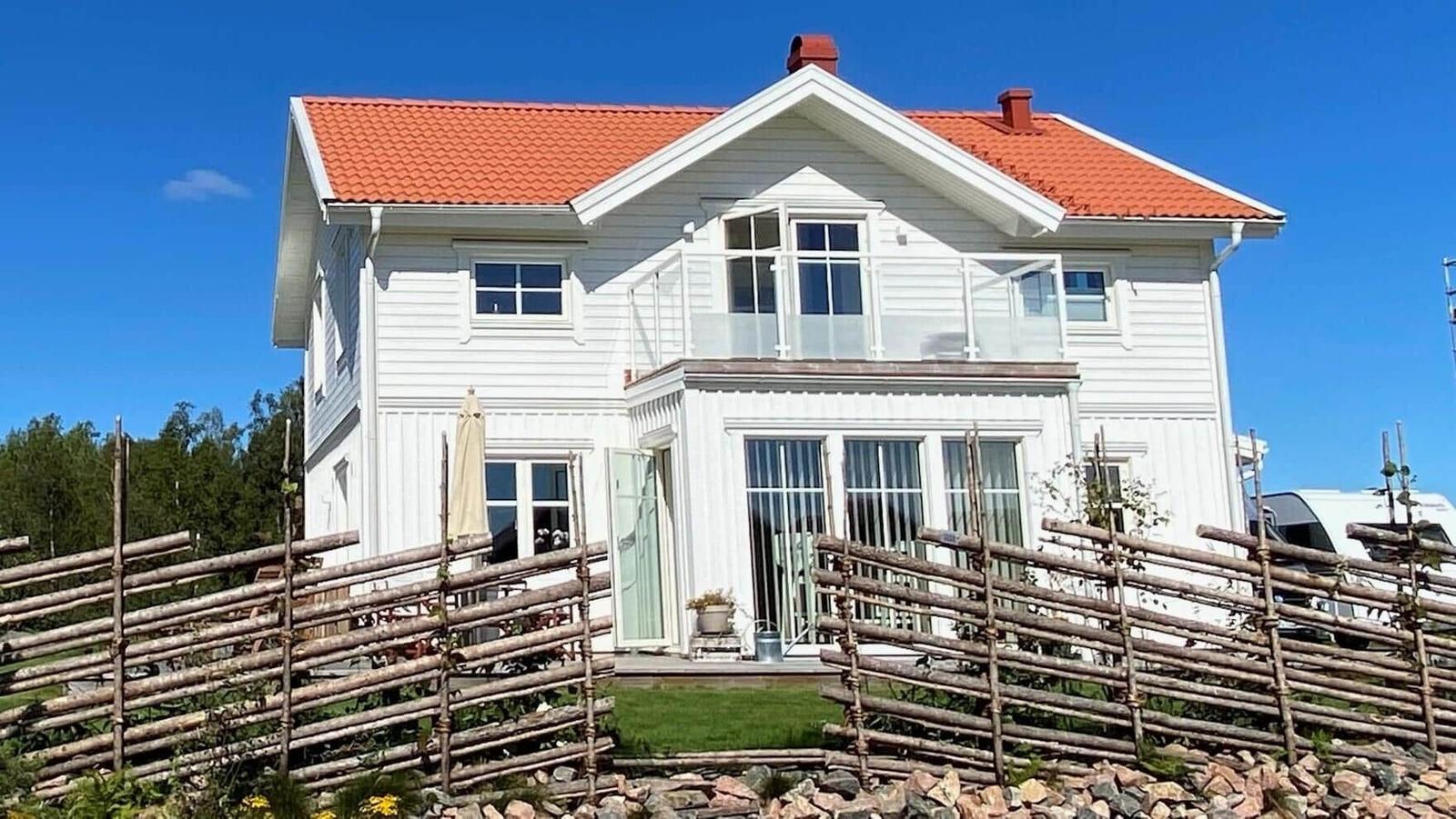 Weiße Villa mit rotem Dach, Balkon und Holzzaun vor grünem Grund.