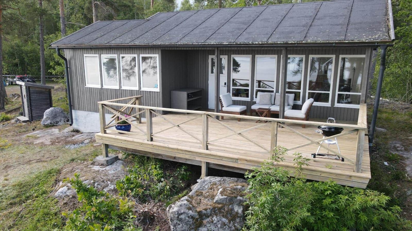 Holzterrasse mit Sitzgelegenheiten und Grill vor einem Haus mit grauer Fassade.