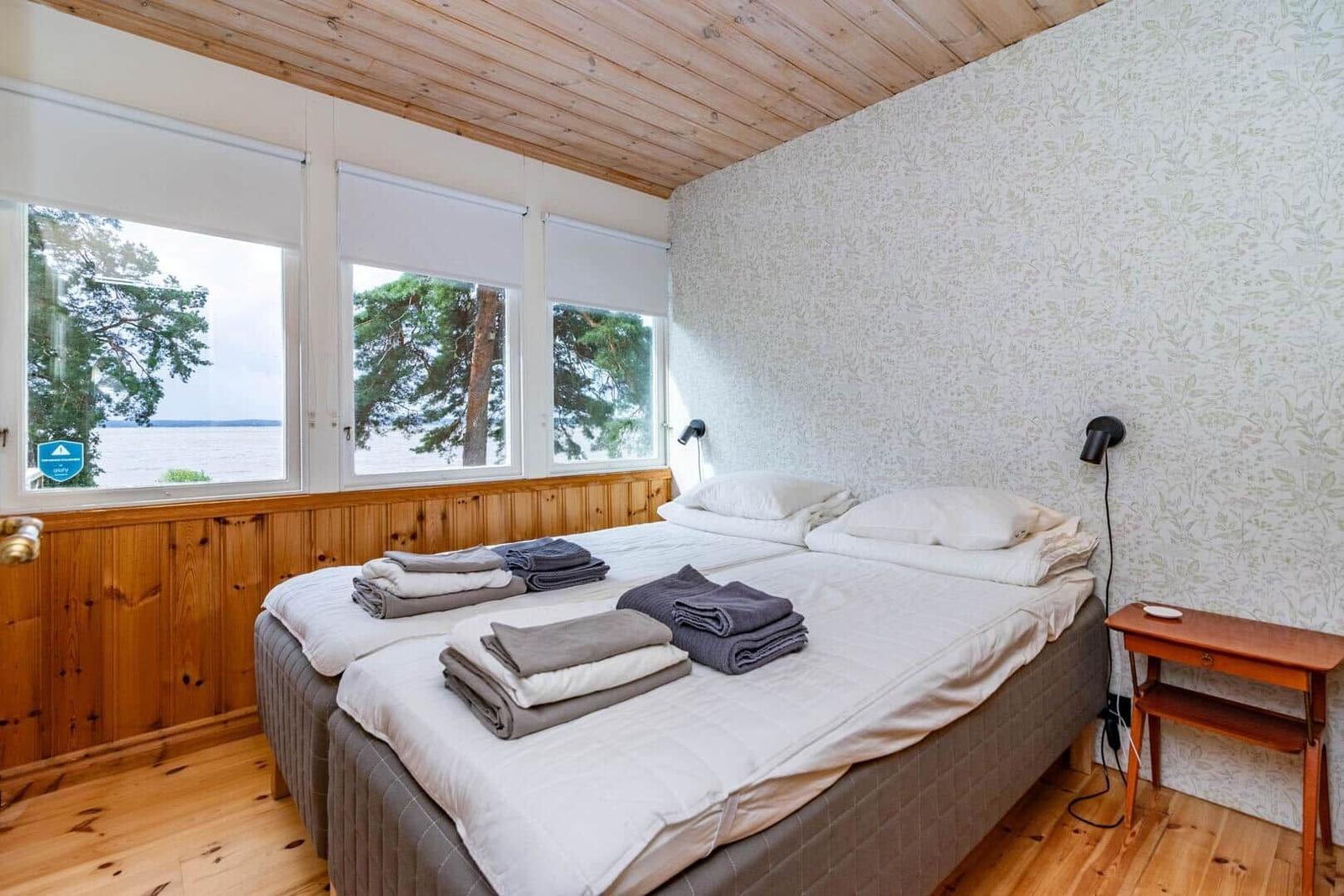 Schlafzimmer mit Doppelbett, Fensterblick auf Wasser und Bäume, Holzdecke und -boden.