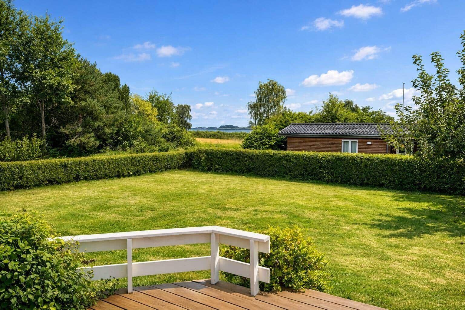 Behind a lawn with hedge stands a wooden house. View of water and trees.