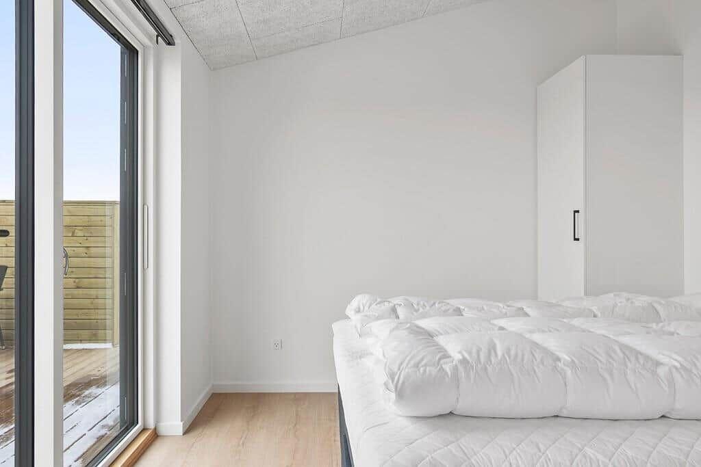 Schlafzimmer mit weißem Bett und großem Fenster zur Terrasse.
