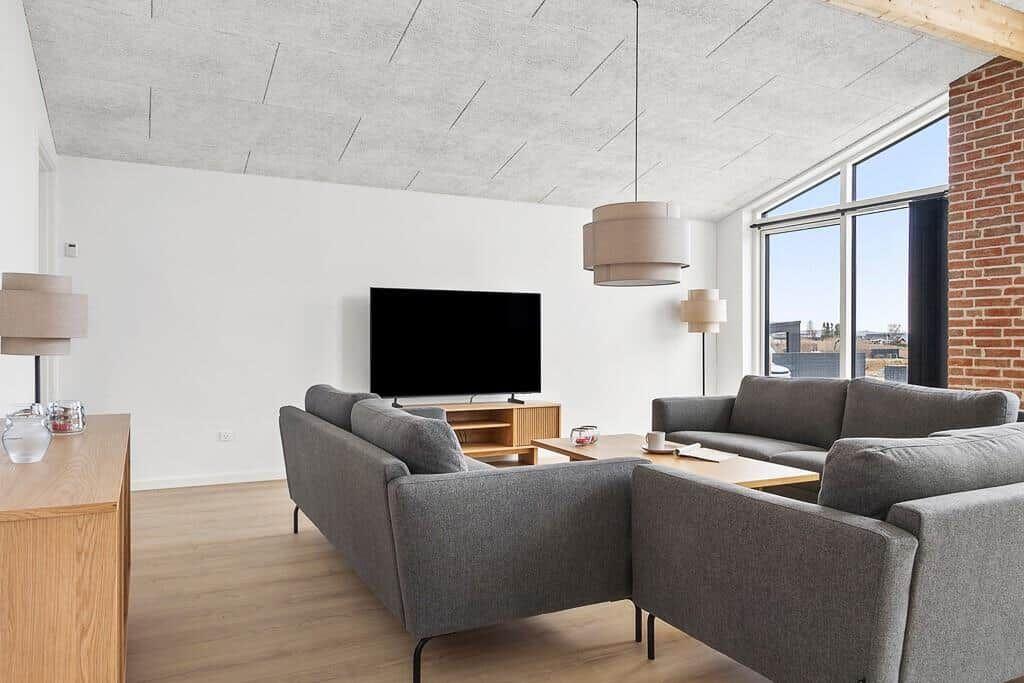 Wohnzimmer mit grauem Sofa, Fernseher und Holzmöbeln. Große Fenster und Backsteinwand.