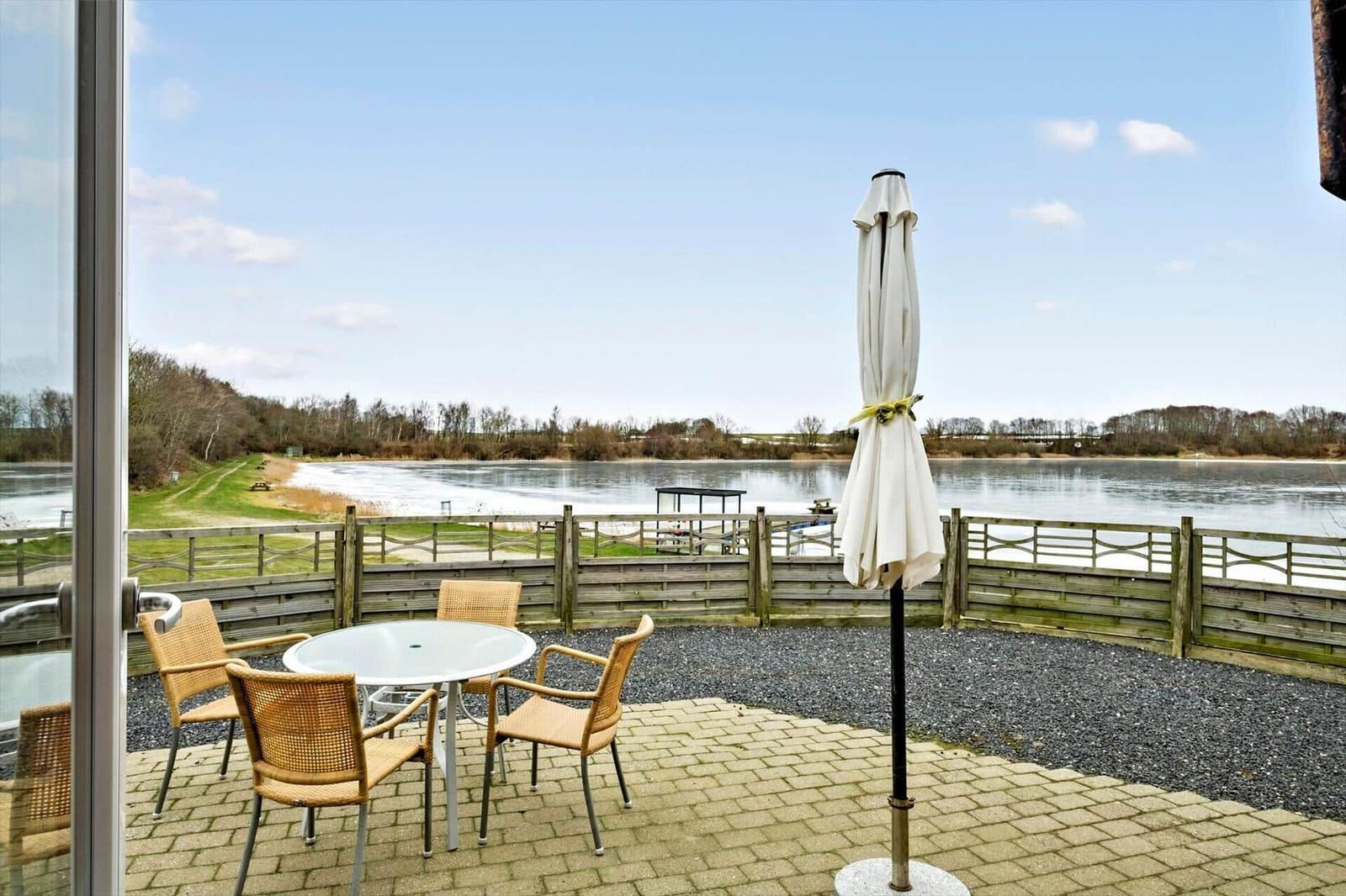 Terrace with table, chairs, and umbrella. View of a lake with shoreline path and woods.
