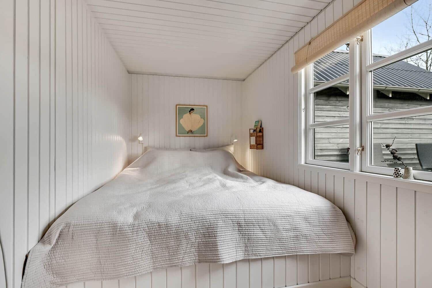 Schlafzimmer mit weißer Holzwandverkleidung, Bett und Fenster mit Blick nach draußen.