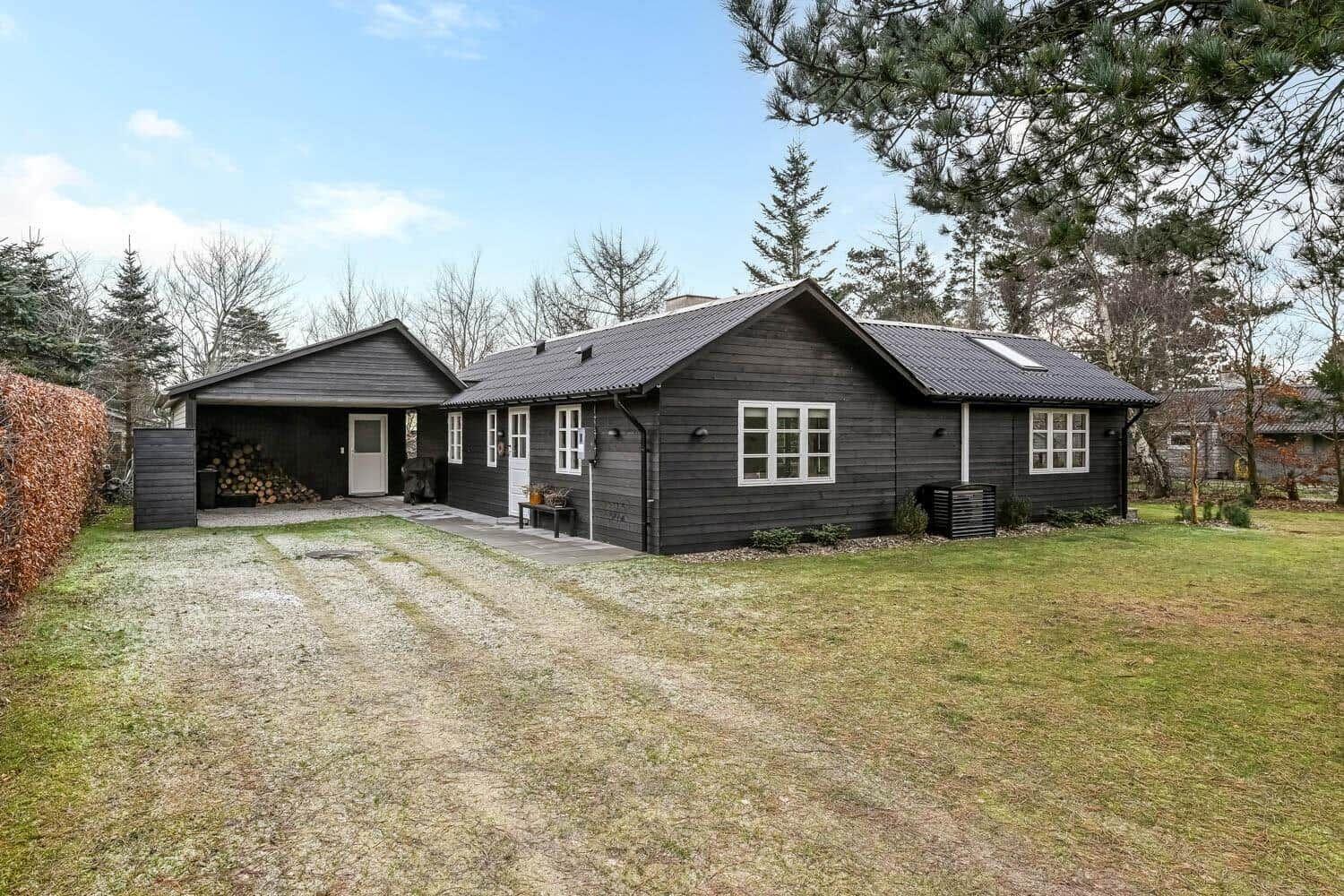 Schwarzes Holzhaus mit Garage und Rasenfläche. Umgeben von Bäumen und Hecke.