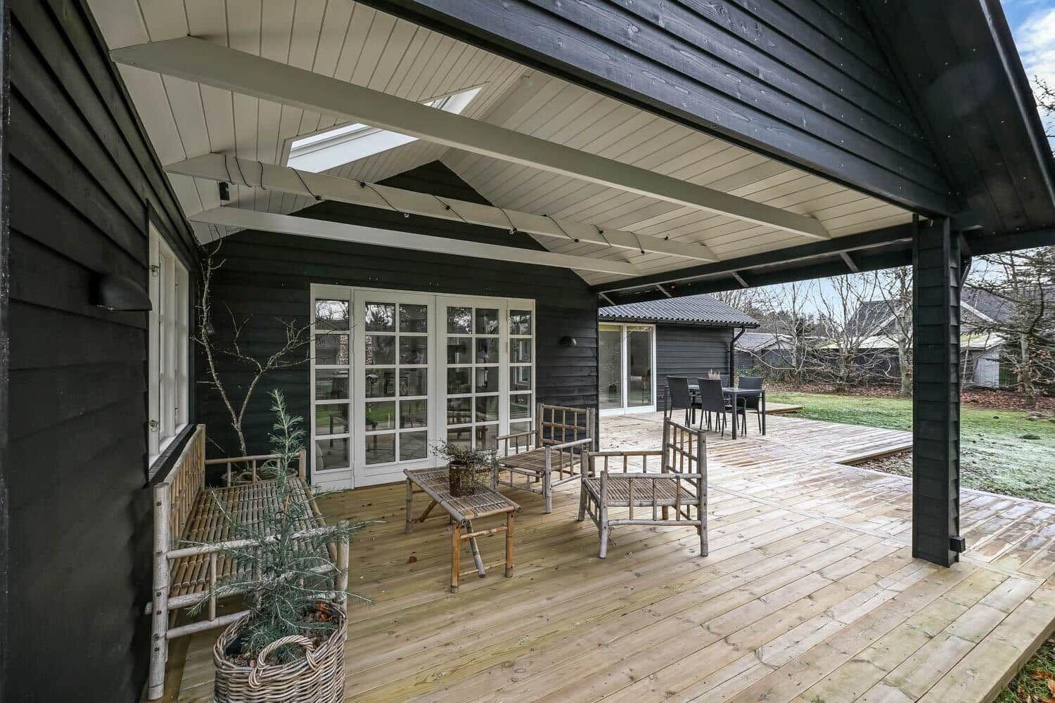Terrasse mit Holzboden, Bänken und Tisch unter Dach. Hintergrund zeigt Garten und Haus.