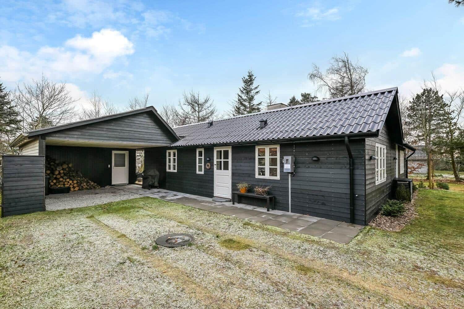 Schwarzes Holzhaus mit weißer Tür und Fenstern, Grillplatz und Holzstapel im Garten.