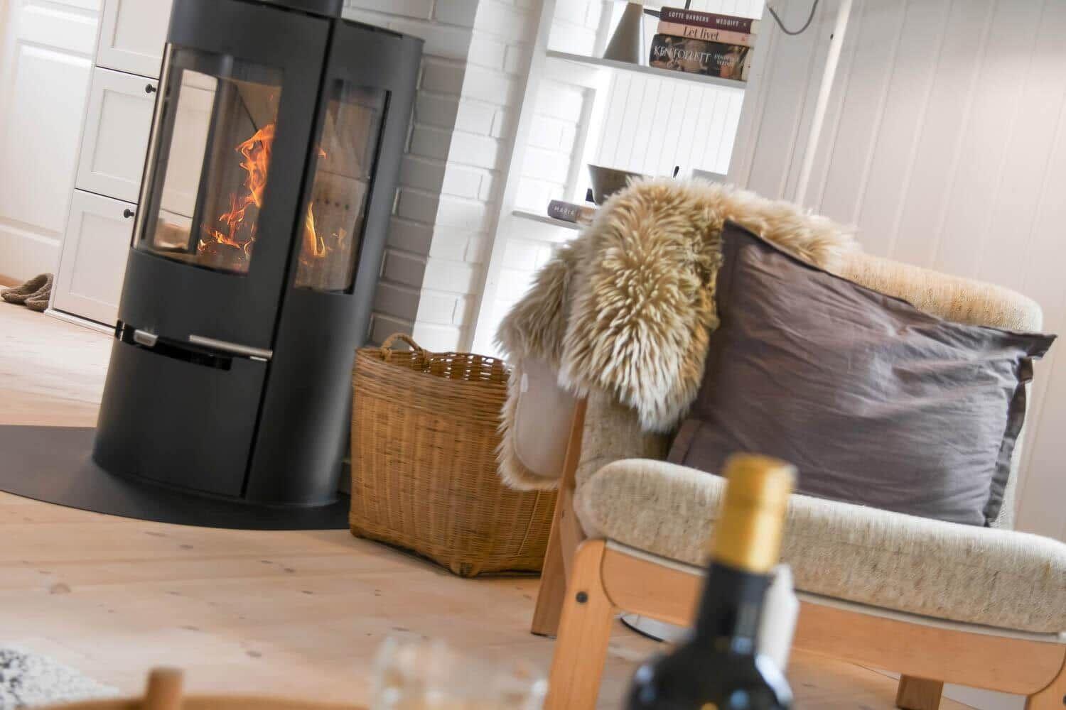 Ein Kamin mit brennendem Holz, ein Sessel mit Felldecke und Kissen, ein Korb und ein Flaschenhals im Vordergrund.