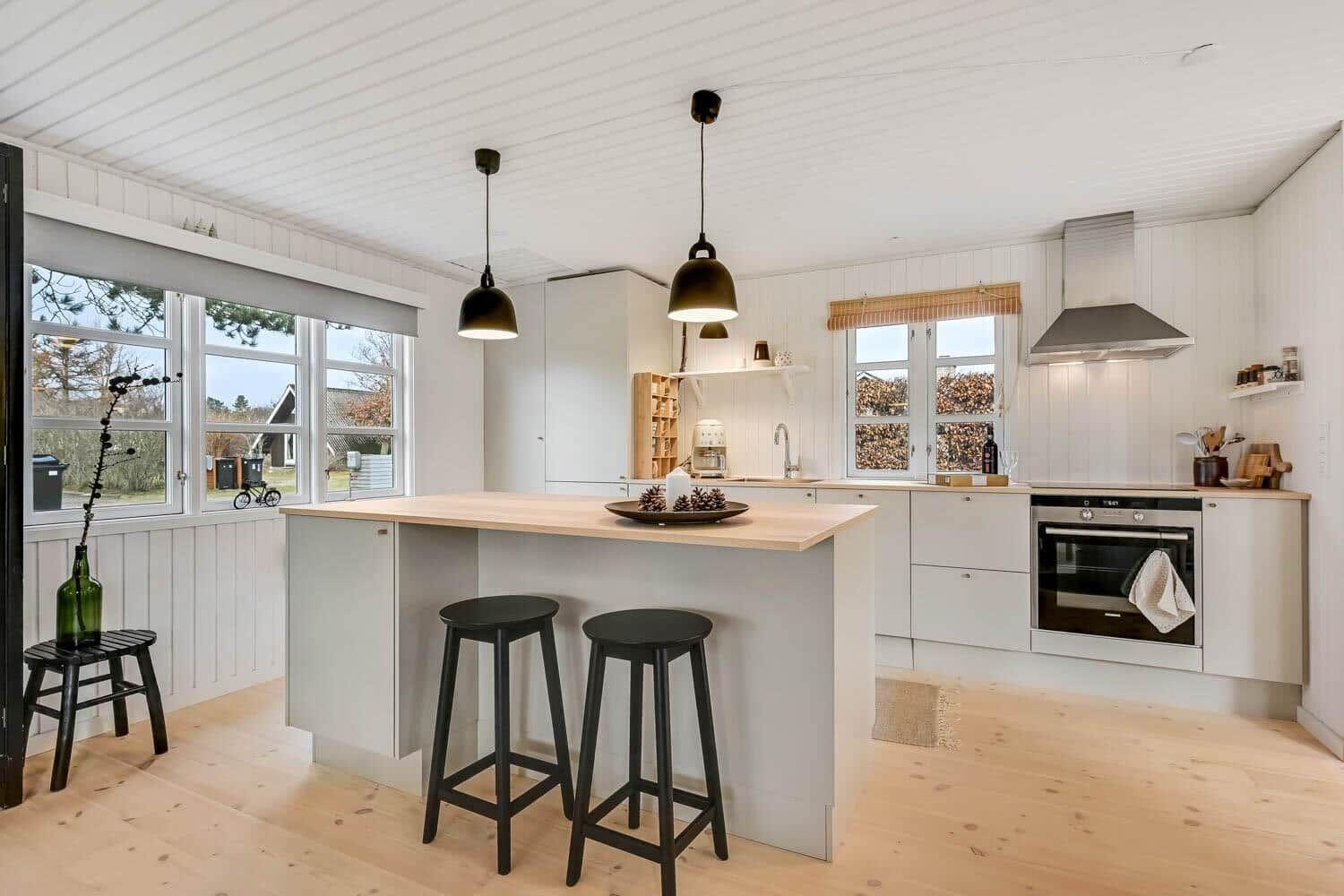 Küche mit Insel, schwarzen Hockern und Holzboden. Weiße Wände und Decke mit Fenstern.