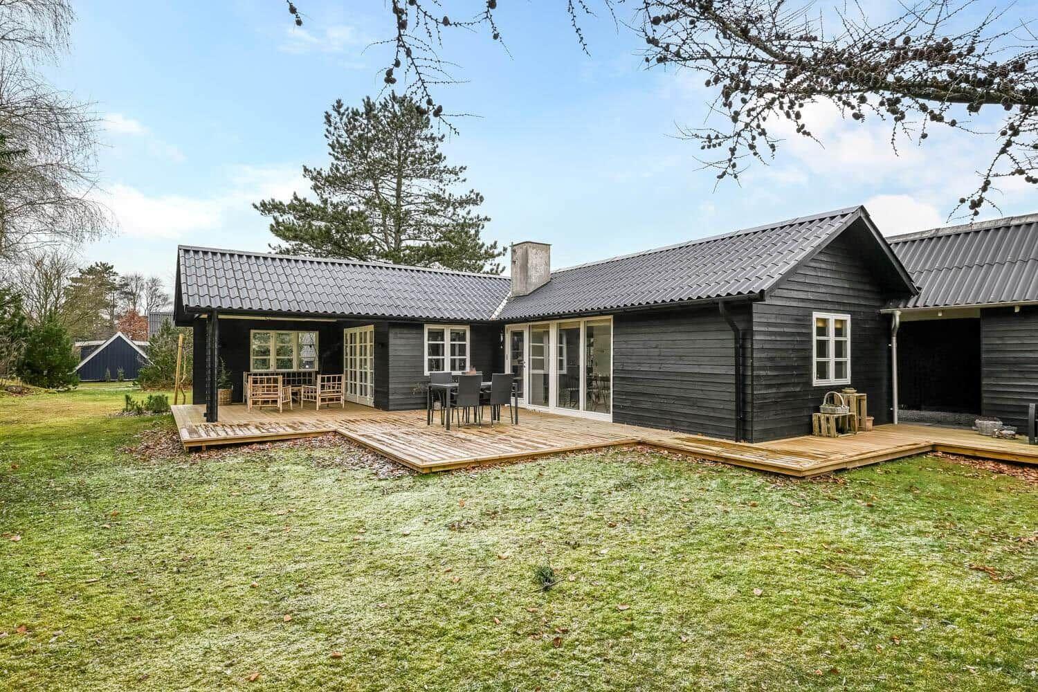 Schwarzes Holzhaus mit Terrasse und Garten. Glasfront und Holzstühle sind sichtbar.