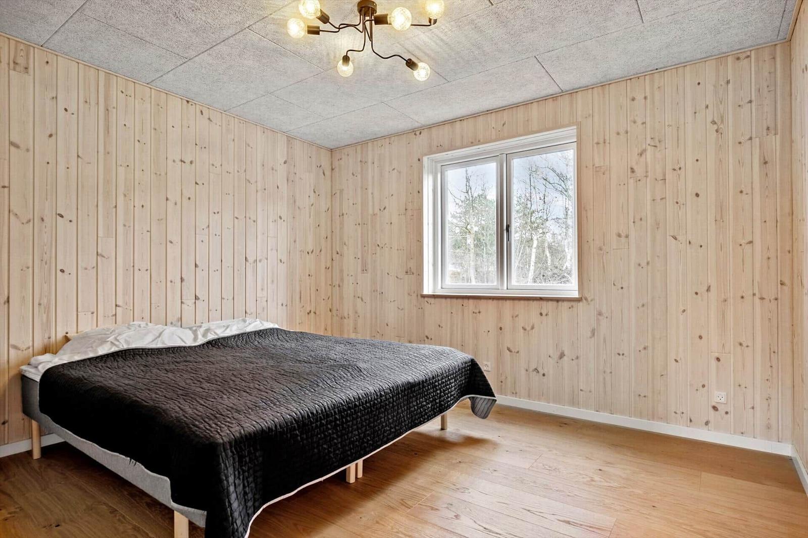 Schlafzimmer mit Holzwand, Bett und Fenster mit Blick auf Bäume.