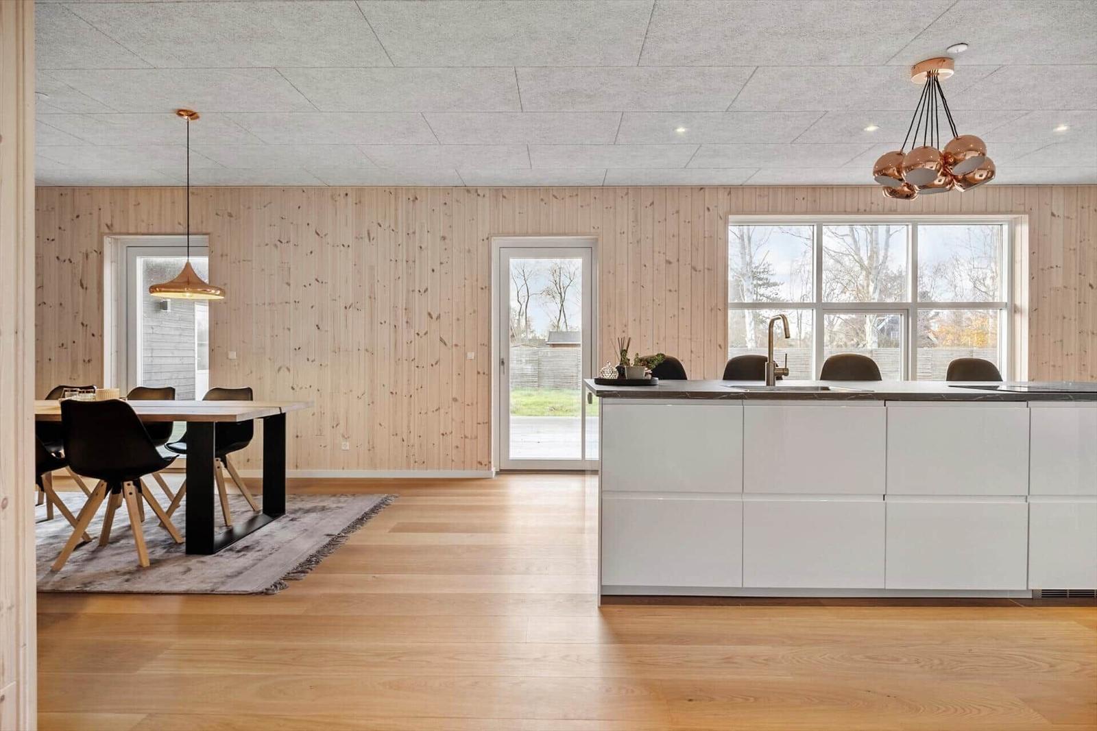 Küche mit Insel und Essbereich. Holzwand, Boden und Fenster sichtbar.