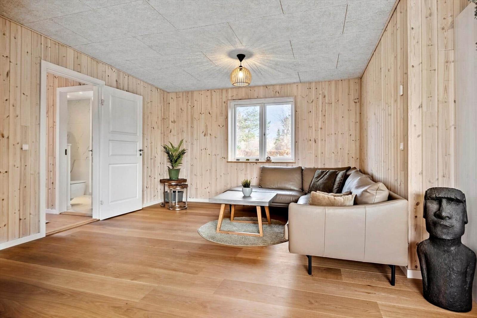 Wohnzimmer mit Holzwand, Sofa, Tisch und Fenster. Tür führt in Bad.