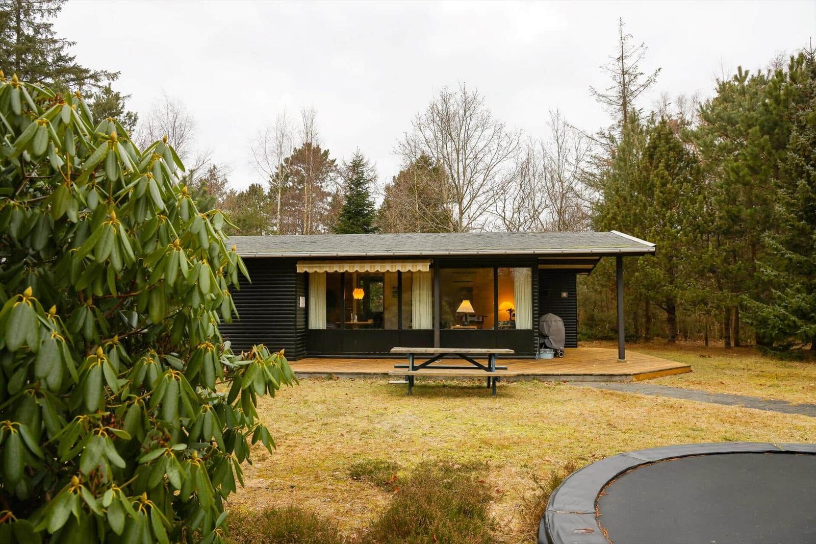 Wooden house with terrace, picnic table, and trampoline in forest area.