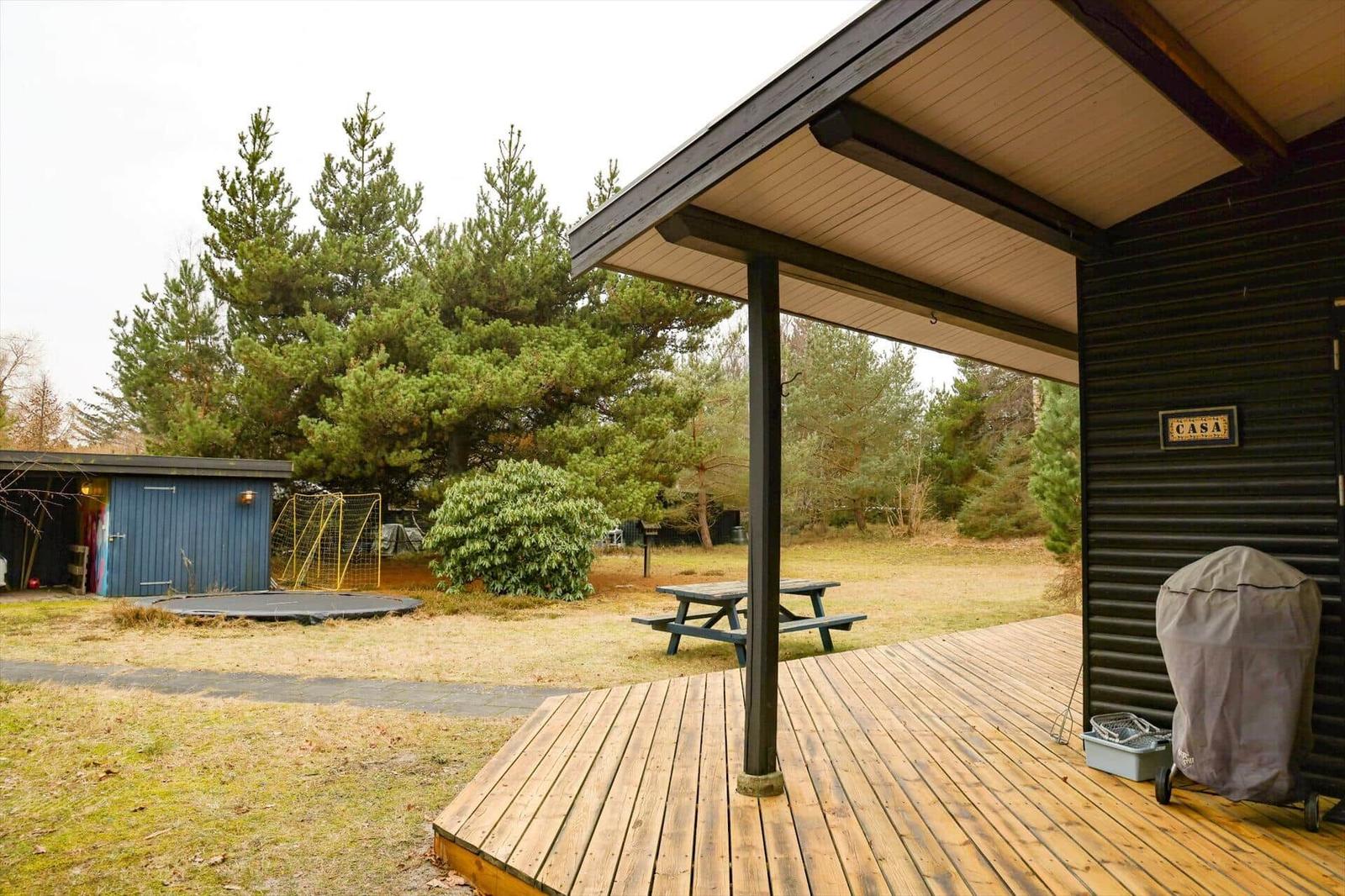 Wooden deck with grill and picnic table in front of a house with garden and trees.