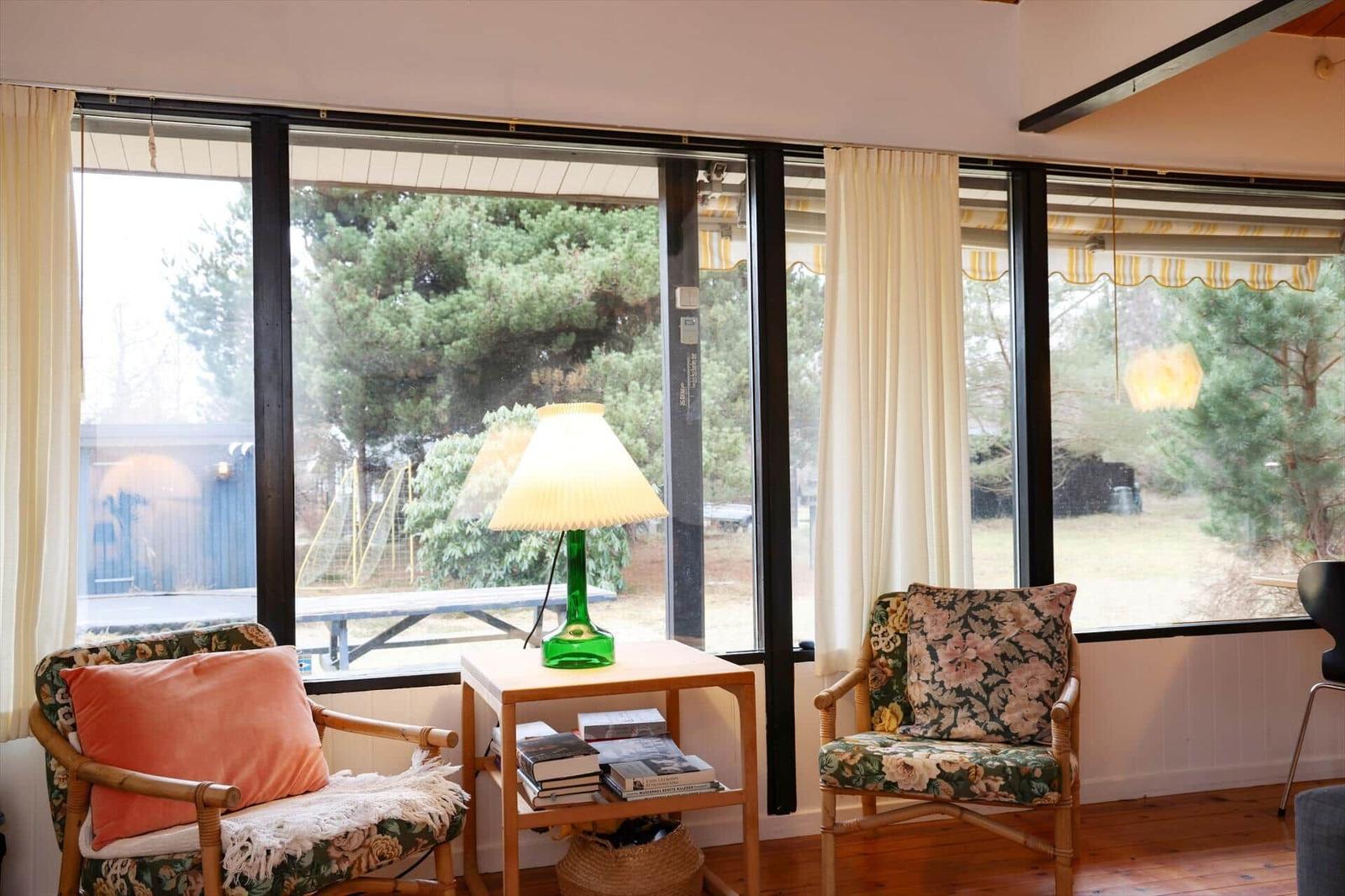 Two chairs with floral upholstery stand before large windows. A green lamp sits on a table.