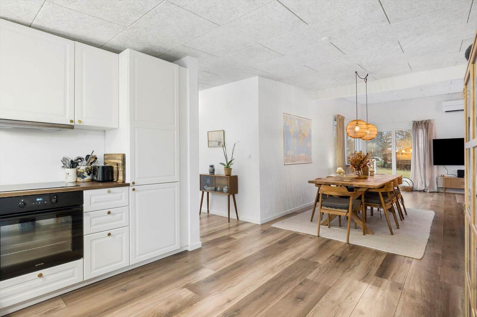 Kitchen with dining area and living room. Wooden floor, white cabinets, dining table with chairs.