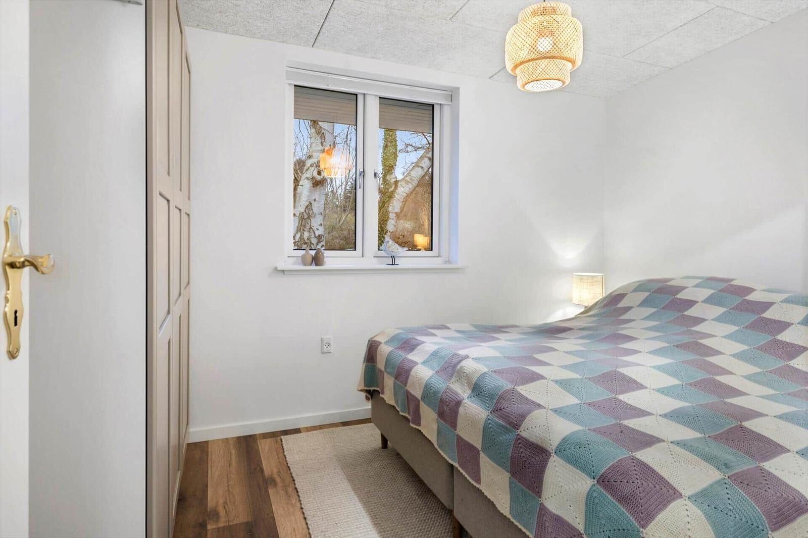 Bedroom with bed, window, and quilt. Wooden floor and lamp.