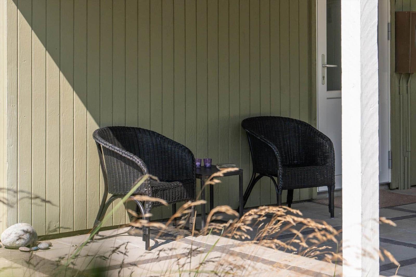 Zwei dunkle Rattan-Stühle stehen auf einer Terrasse vor einer grünen Holzwand.