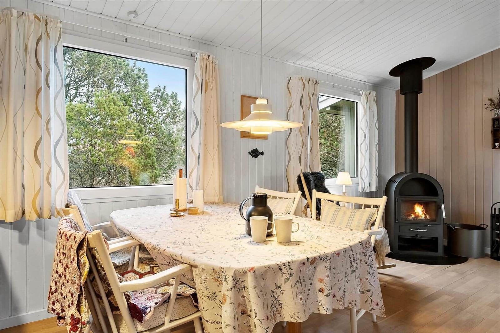 Esszimmer mit Tisch, Stühlen und Kaminofen. Große Fenster zeigen grüne Bäume.