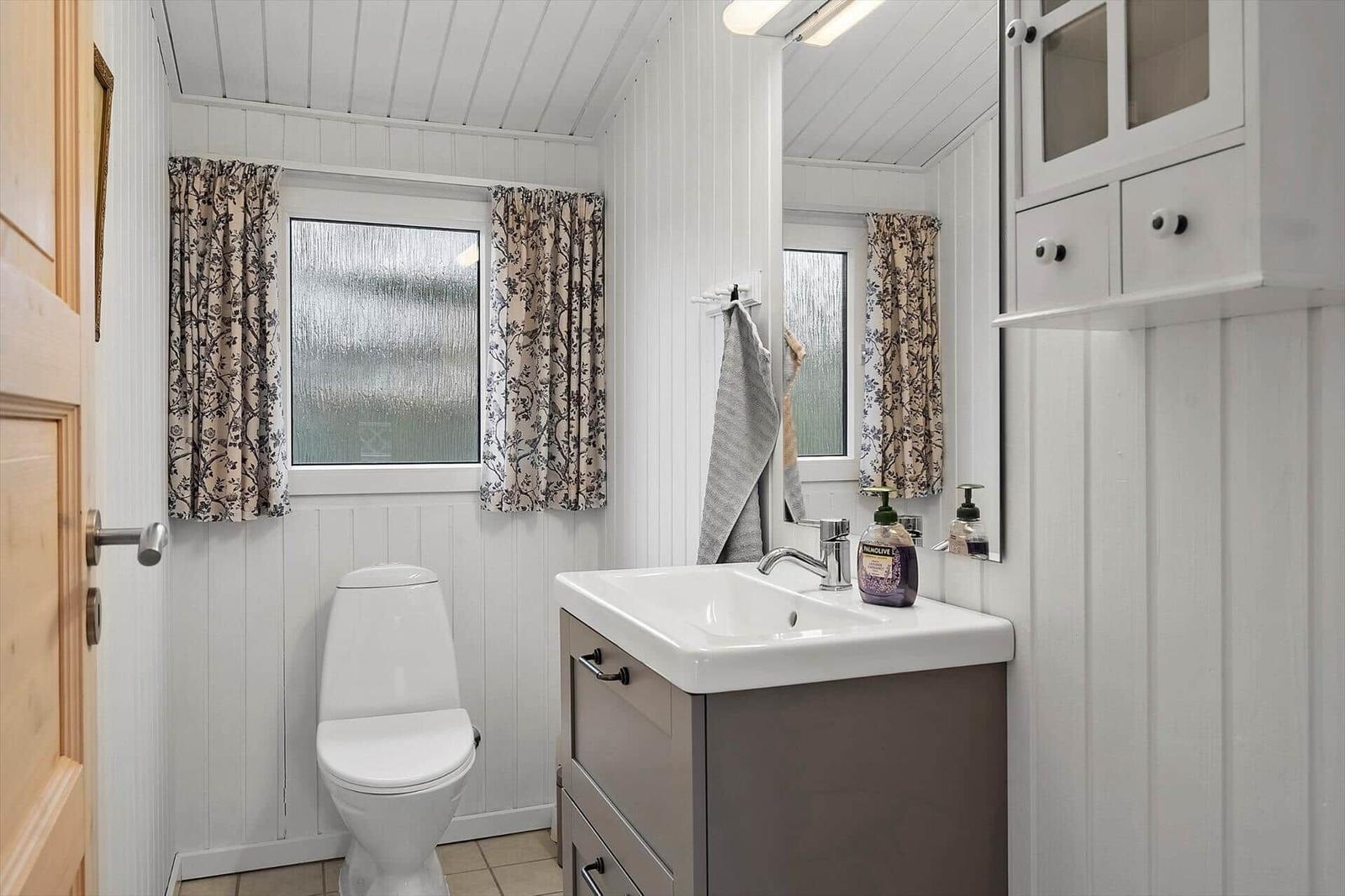 Badezimmer mit Toilette, Waschbecken und Spiegel. Fenster mit Vorhängen.