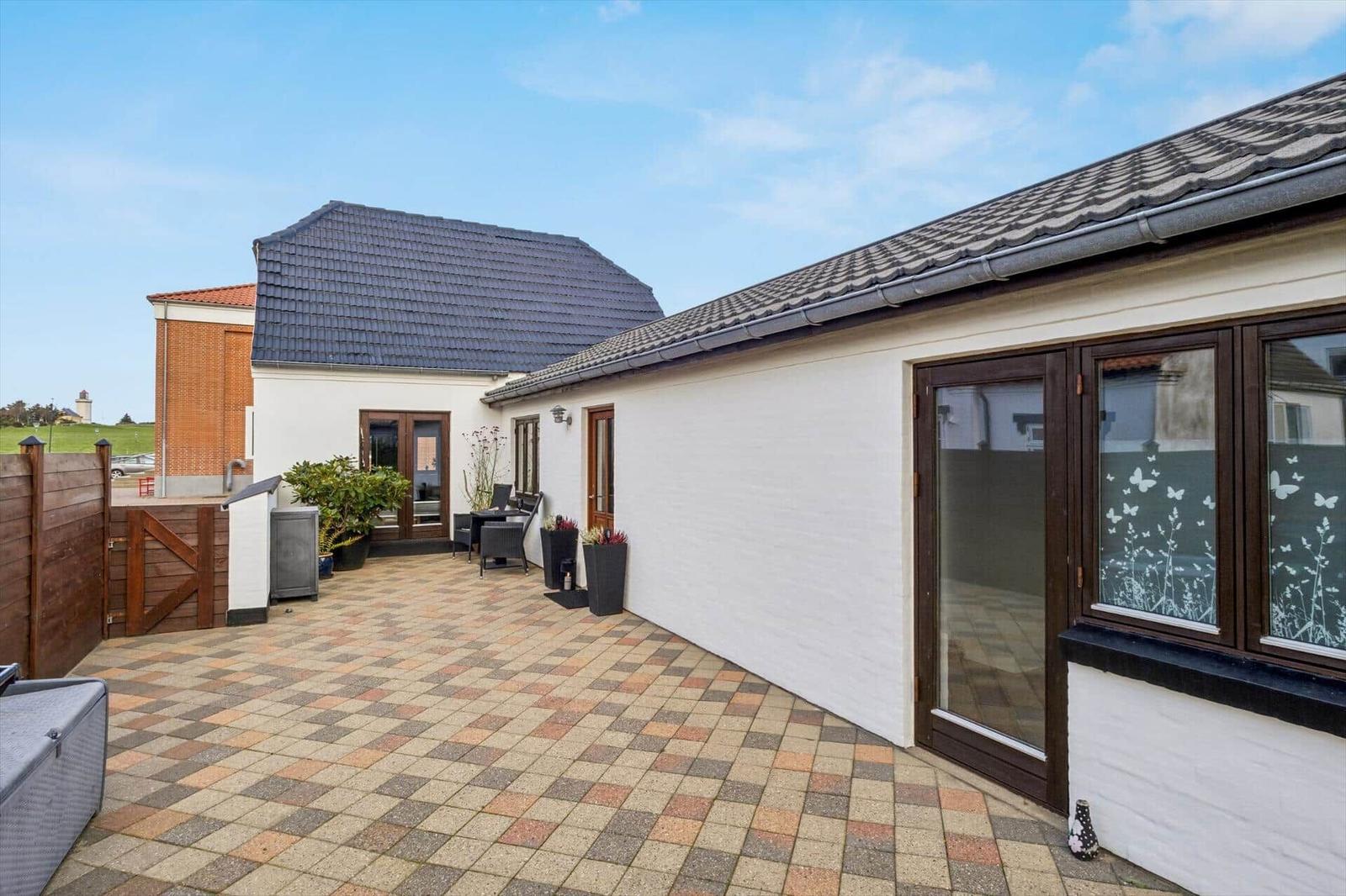 Backyard with terrace, garden furniture, and plants. White wall, brown windows, and roof tiles.