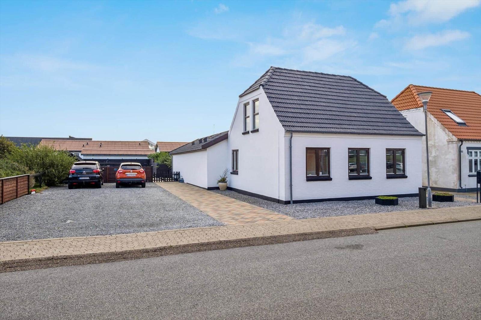 White house with dark roof, paved path, and gravel area. Two cars parked in front of the house.