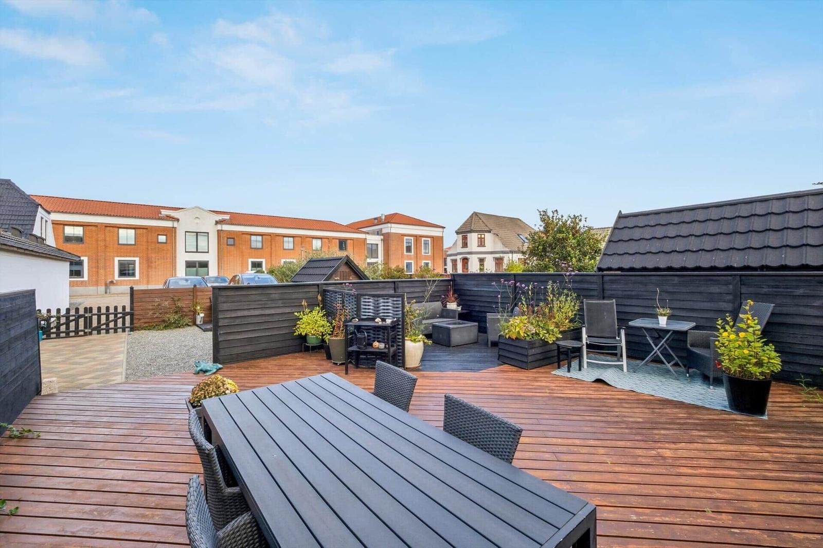 Deck with wooden flooring, seating area, and plants. View of residential buildings in the background.