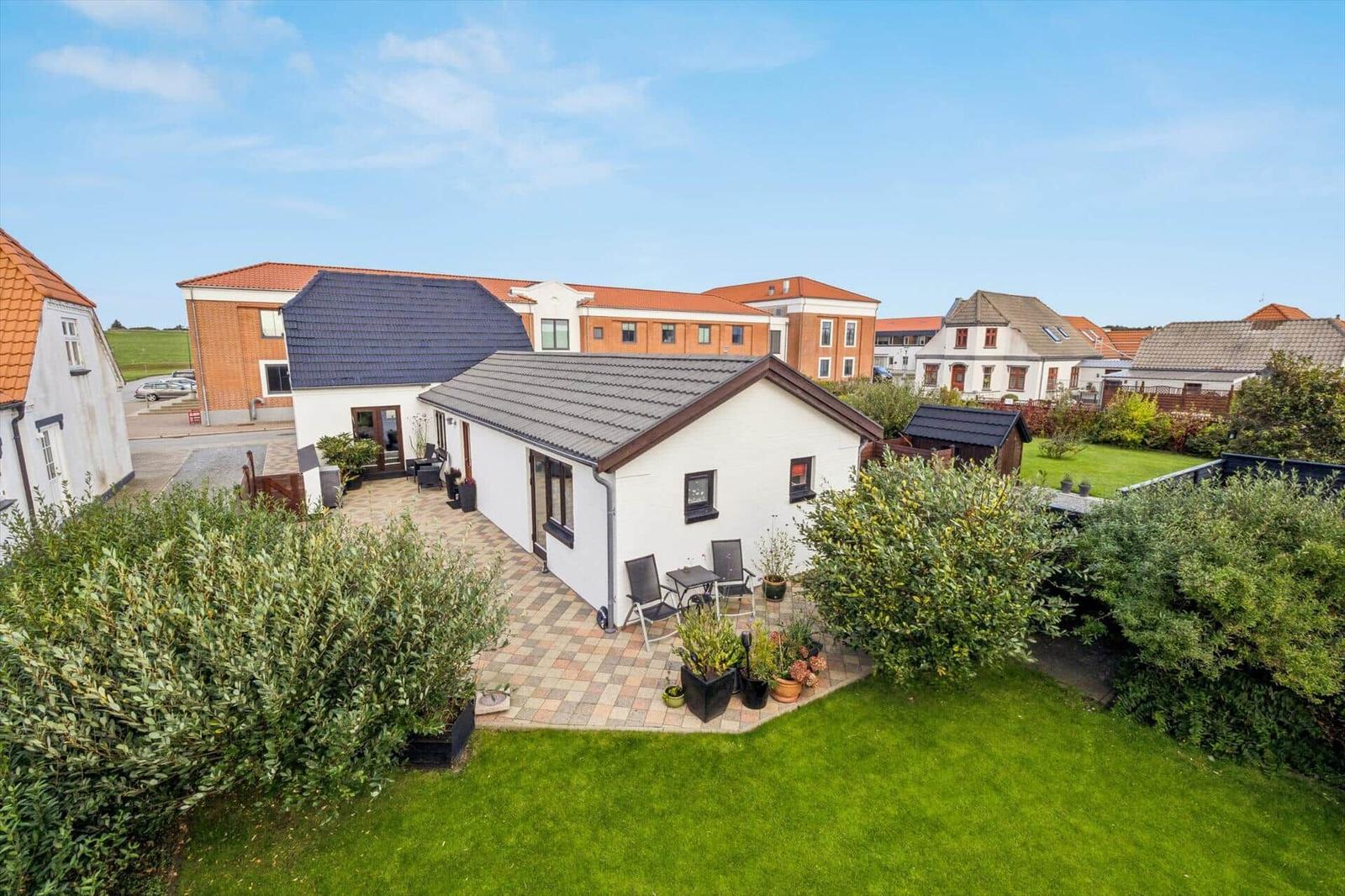 White house with terrace and garden. Surrounded by greenery and neighboring houses.