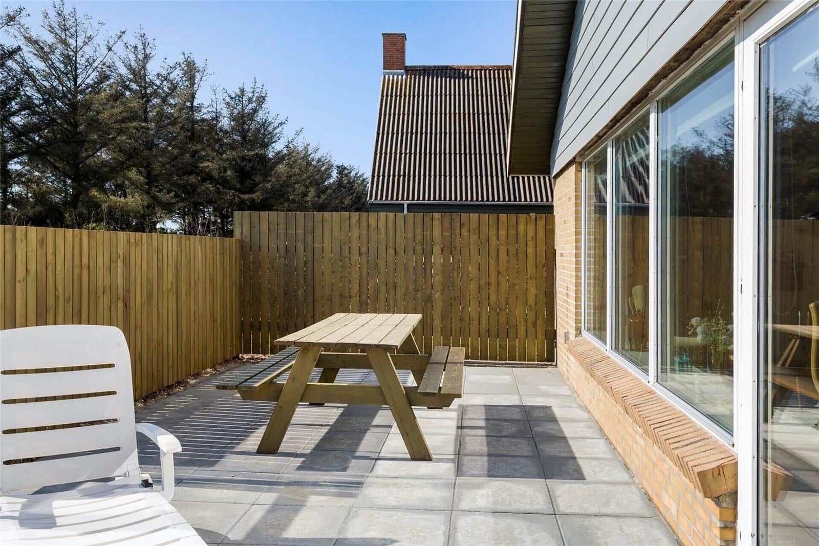 Terrasse mit Picknicktisch, weißem Stuhl und Holzzaun. Blick auf Haus mit Schiebetür.