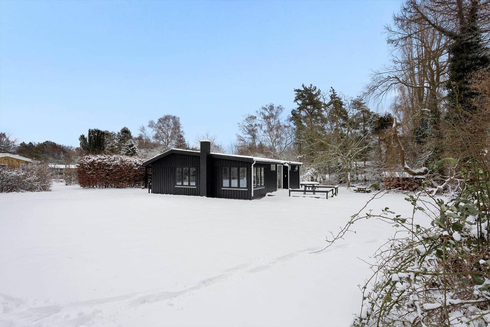 Black wooden house with snow-covered ground and trees in the background.