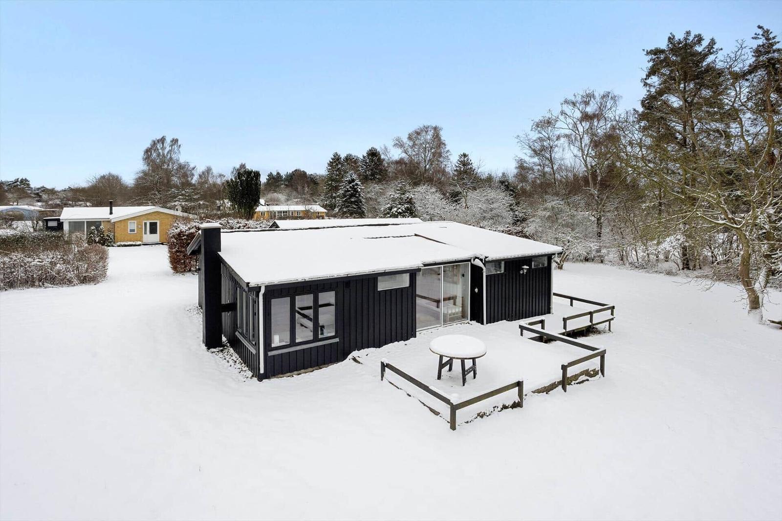 Black-painted cabin with snow-covered roof and terrace in winter.