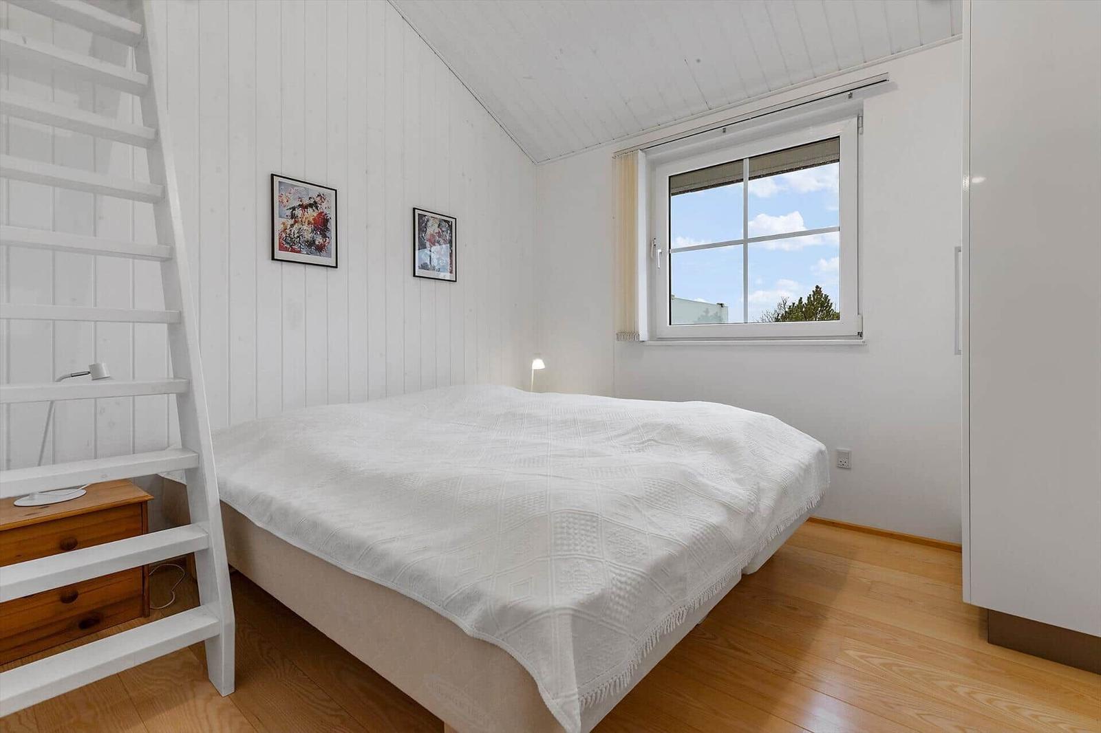 Schlafzimmer mit weißem Bett, Fenster und Holzboden. Links Leiter und Nachttisch.