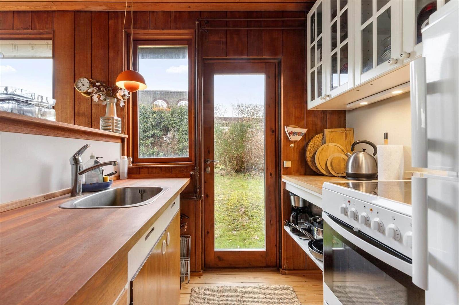 Küche mit Holzwand, Fenster und Tür zum Garten. Weiße Schränke, Edelstahlkochtopf und Arbeitsfläche.