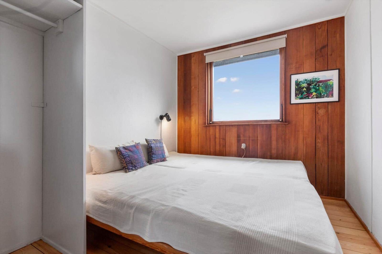 Schlafzimmer mit Bett, Fenster und Holzwand. Kleine Nachttischlampe und Bild an der Wand.