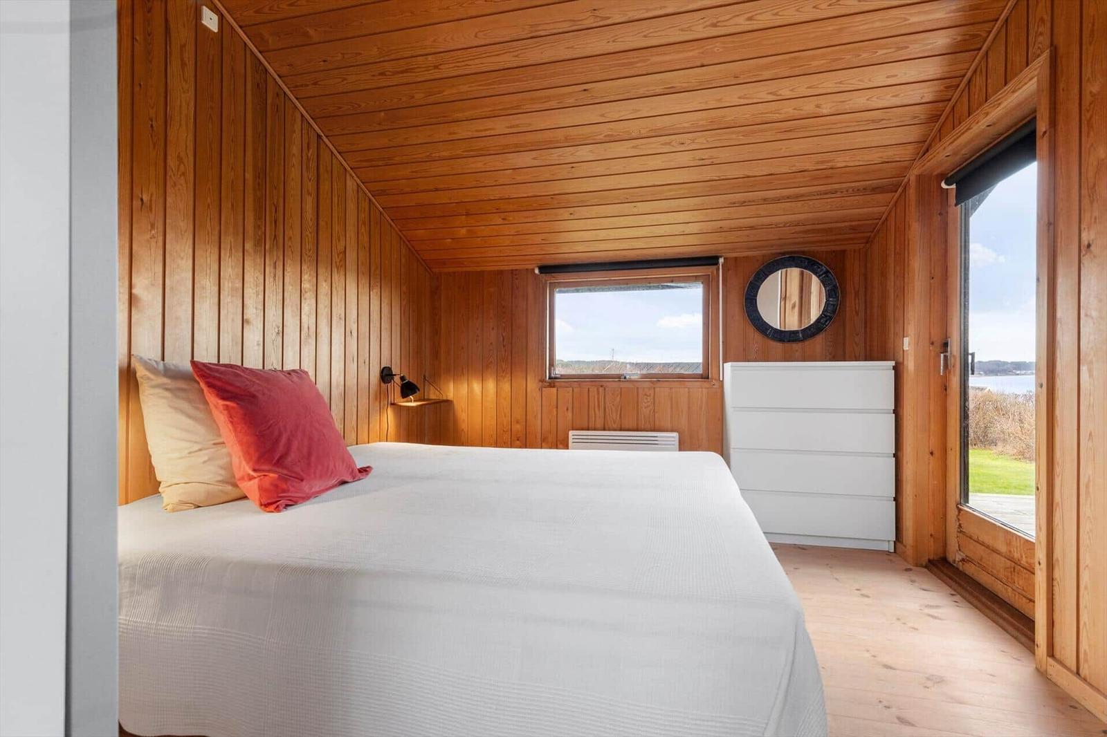 Schlafzimmer mit Holzwand, Bett, Schrank und Fensterblick auf Wasser