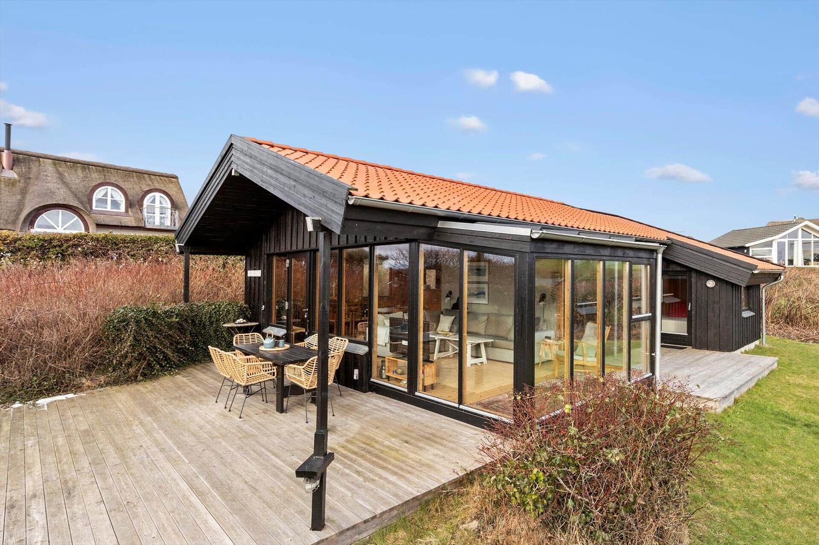 Moderne Holzhütte mit Glasfront, Terrasse und Garten. Sichtbarer Innenraum mit Möbeln.