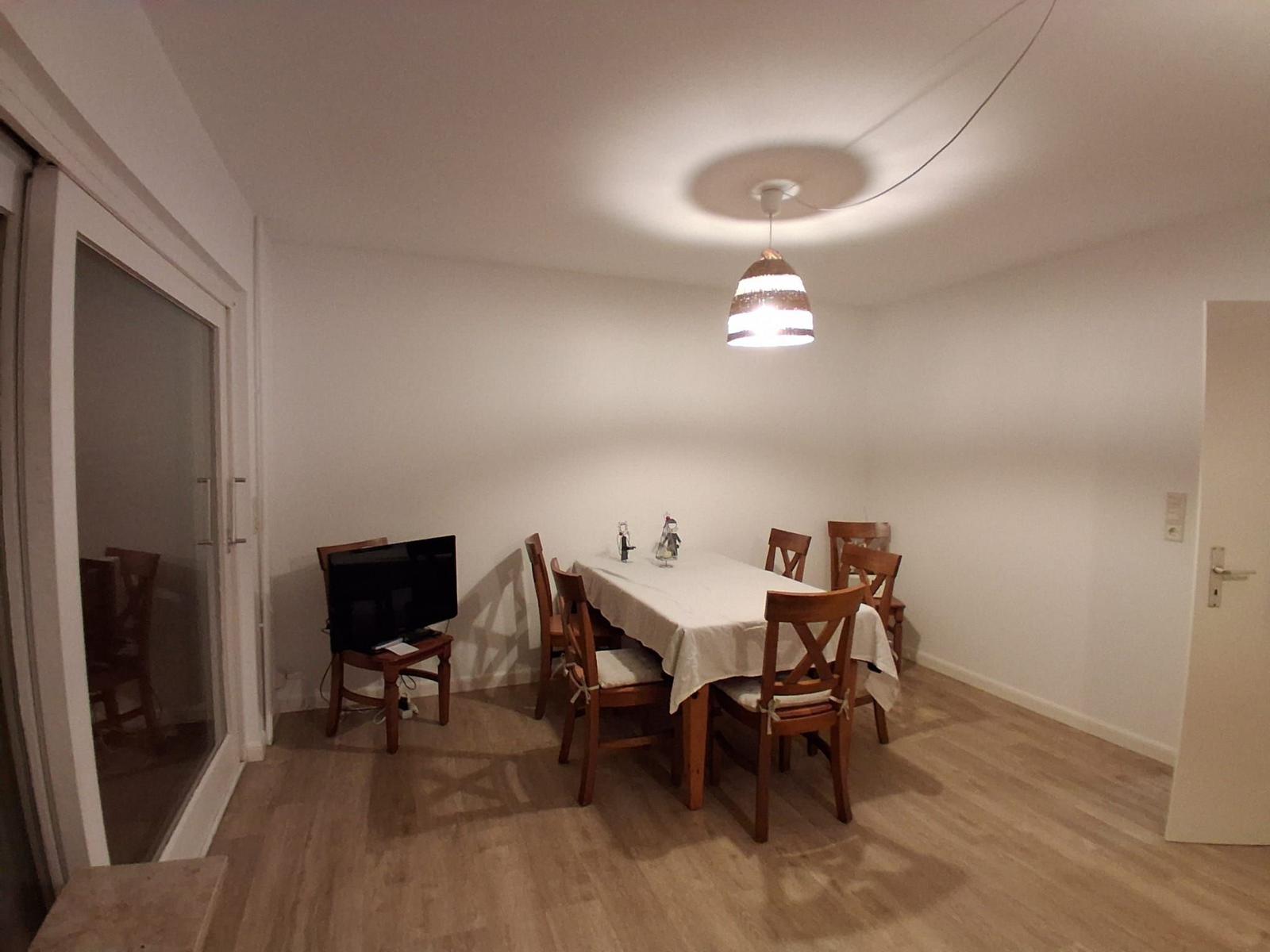 Esszimmer mit Holztisch, Stühlen und Deckenleuchte. Fernseher und Glastür sichtbar.