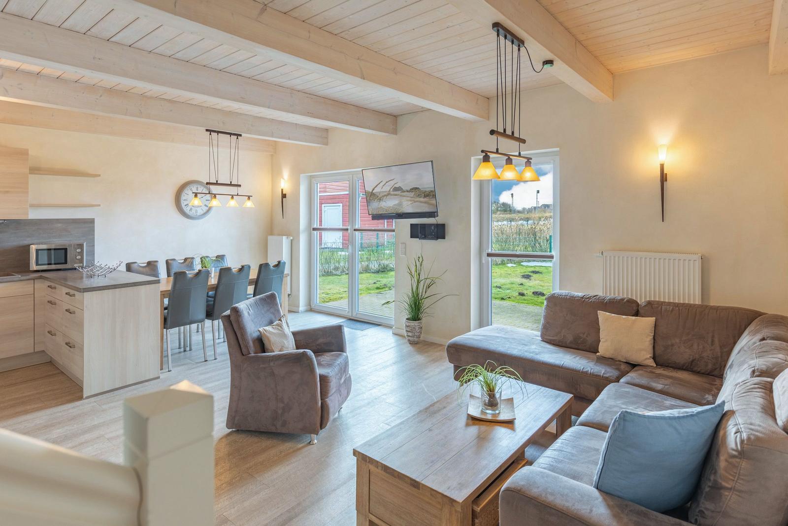 Wohn- und Essbereich mit Sofa, Sessel und Küchenzeile. Große Fenster mit Blick auf Grünfläche.