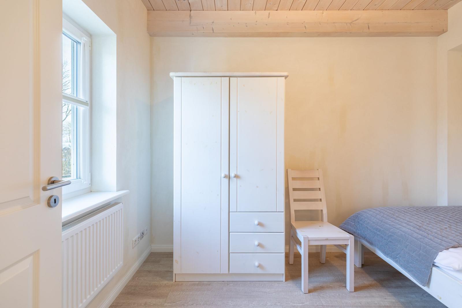 Schlafzimmer mit Bett, Kleiderschrank und Stuhl. Holzdecke und Fenster mit Heizkörper.