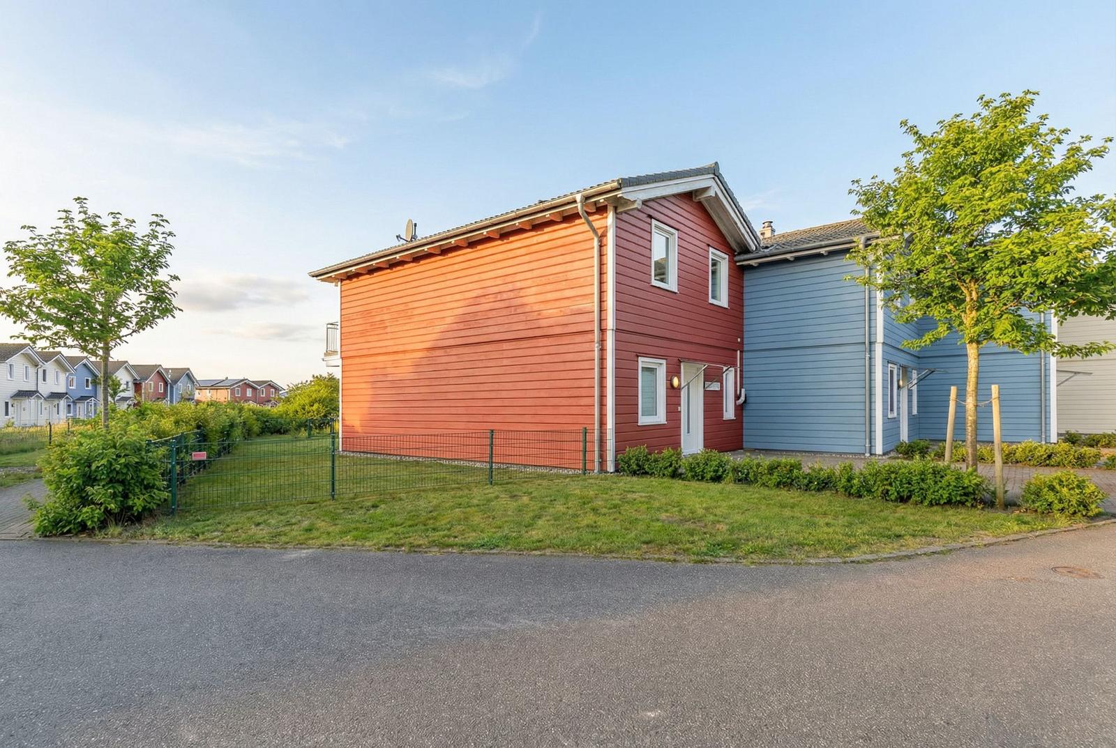 Rotes und blaues Haus mit Garten und Zaun. Hintergrund: weitere Häuser.