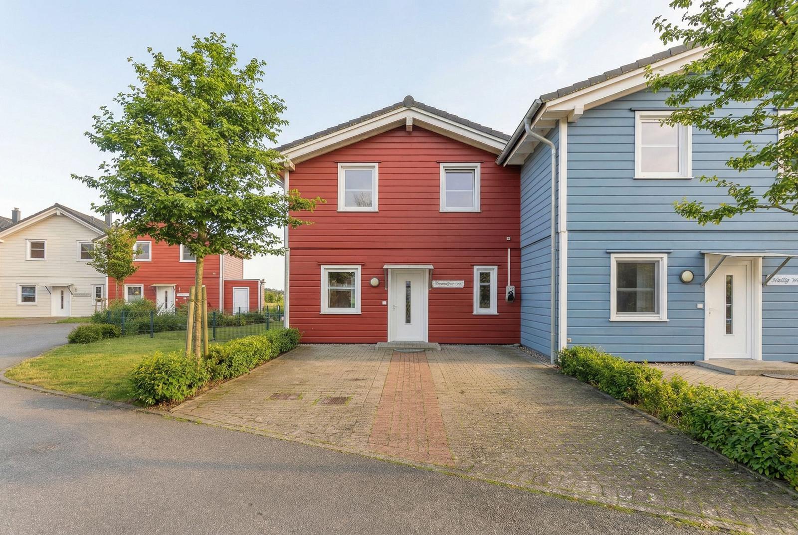 Rotes und blaues Ferienhaus mit gepflastertem Vorplatz und grüner Umgebung.
